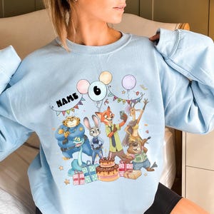 May include: Pale blue sweatshirt with a vibrant cartoon design of characters, balloons, and a birthday cake. The word "NAME" is printed above the characters. The number "6" is displayed on a balloon. Presents and stars are also included in the design.