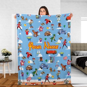 May include: A light blue blanket featuring cartoon characters and the text "Your Name". The blanket has a repeating pattern of various characters, cars, and clouds. The blanket is a soft, cozy item.