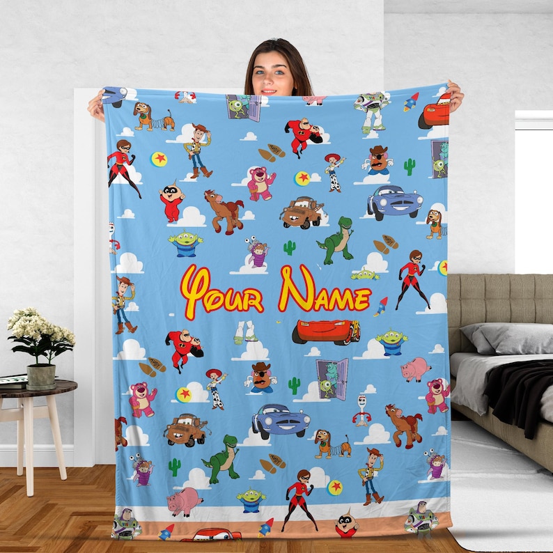 May include: A light blue blanket featuring cartoon characters and the text "Your Name". The blanket has a repeating pattern of various characters, cars, and clouds. The blanket is a soft, cozy item.