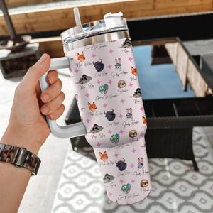 Puede incluir: Un vaso de color rosa claro con asa y tapa blancas, con un patr&oacute;n repetido de personajes de dibujos animados y texto. El vaso incluye una pajita y est&aacute; sujeto por una mano. El texto incluye las frases "Gary Di Snake", "Judy Hopper" y "Mr. Big".