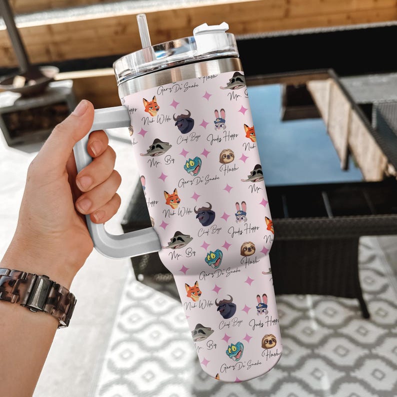 Puede incluir: Un vaso de color rosa claro con asa y tapa blancas, con un patr&oacute;n repetido de personajes de dibujos animados y texto. El vaso incluye una pajita y est&aacute; sujeto por una mano. El texto incluye las frases "Gary Di Snake", "Judy Hopper" y "Mr. Big".