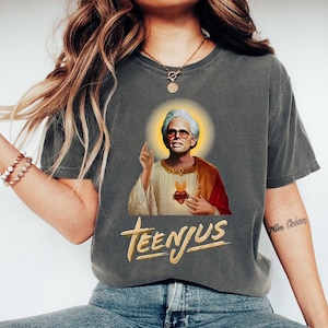 Pode incluir: T-shirt cinzento escuro com um gráfico de um homem com uma auréola, óculos de sol e uma túnica vermelha e branca. O texto "TEENJUS" está impresso em dourado sob a imagem. A camisa é de estilo casual.