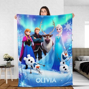 Puede incluir: Una manta polar azul con personajes de la pel&iacute;cula animada Frozen, incluyendo a Anna, Elsa, Kristoff, Sven y Olaf. La manta tiene un fondo invernal con copos de nieve y el nombre "OLIVIA" en la parte inferior.