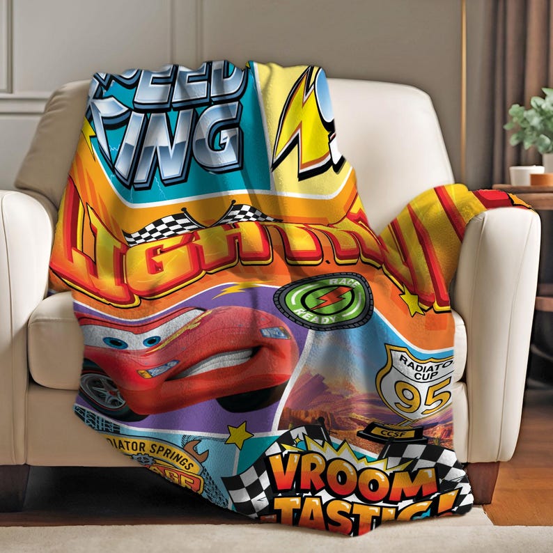 May include: A colorful blanket featuring Lightning McQueen from the movie Cars. The blanket has a vibrant design with the words "SPEED WING LIGHTNING" and "VROOM TASTIC!" printed on it. The blanket is draped over a white armchair.