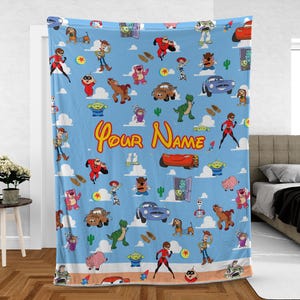 May include: A light blue blanket featuring a repeating pattern of cartoon characters, including Woody, Buzz Lightyear, and others. The blanket has the text "Your Name" in a yellow script font. The blanket is displayed in a bedroom setting.
