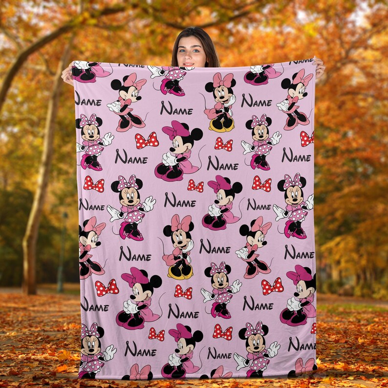 May include: A pink blanket with a repeating pattern of Minnie Mouse characters, bows, and the word "Name". The blanket has a soft, plush appearance, suitable for a child's room or as a gift.