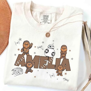 Può includere: T-shirt color crema con un design personalizzato con il nome "AMELIA" in marrone, insieme a illustrazioni di Chewbacca, BB-8 e astronavi. La maglietta ha un girocollo e maniche corte. Una collana d'oro con un ciondolo è drappeggiata sulla maglietta.
