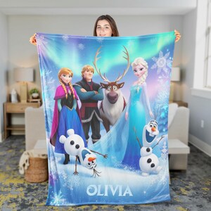 Puede incluir: Una manta de forro polar azul y blanco con personajes de la pel&iacute;cula Frozen, incluyendo a Elsa, Anna, Kristoff, Sven y Olaf. La manta tiene el nombre personalizado "OLIVIA" en la parte inferior.