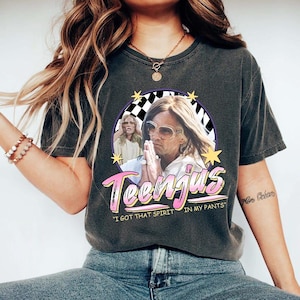 Pode incluir: T-shirt cinza escuro com um gráfico de um homem com óculos de sol e a palavra "Teenjus" em rosa e branco. A camisa também inclui a frase "I GOT THAT SPIRIT IN MY PANTS".