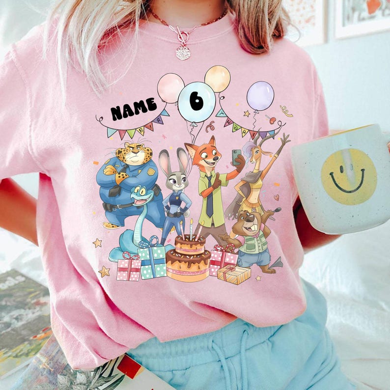 May include: Pale pink sweatshirt with a cartoon graphic of characters, balloons, and a birthday cake. The number "6" is shown, along with the word "NAME" on a banner. A mug with a smiley face is held in the hand.