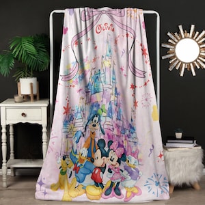 Puede incluir: Una manta rosa con una ilustración de acuarela de un castillo y personajes de Disney, incluyendo Mickey y Minnie Mouse, Donald Duck y Goofy. La manta tiene una cinta con el nombre "Olivia". La manta se exhibe en un estante blanco.