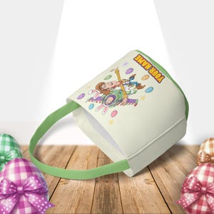Puede incluir: Una cesta de Pascua de color crema con asa y ribete verdes. La cesta presenta una imagen de dibujos animados de Woody y Buzz Lightyear de Toy Story, con el texto "YOUR NAME". Huevos de Pascua decorativos est&aacute;n dispersos alrededor de los personajes.