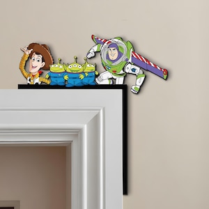 Puede incluir: Un marco de puerta decorativo con personajes de la película de animación Toy Story. Woody, los extraterrestres y Buzz Lightyear están representados en colores vibrantes. Las figuras están posicionadas como si estuvieran encima de un marco de puerta.
