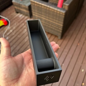 May include: A dark gray rectangular 3D-printed object with a hollow interior and a cylindrical support. The object has a geometric design on one end. The item is held in a person's hand.