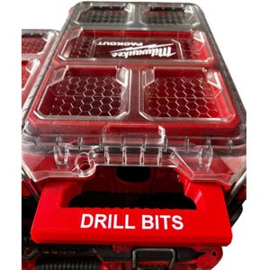 Puede incluir: Un contenedor de almacenamiento de brocas Milwaukee Packout de plástico rojo y transparente. El contenedor tiene un asa roja con las palabras "DRILL BITS" en blanco. La parte superior del contenedor es transparente con detalles en rojo.
