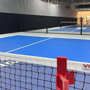 May include: Indoor pickleball court with a blue playing surface and white lines. A red net post is attached to the net. The court is in a large indoor facility with high ceilings and metal support beams.