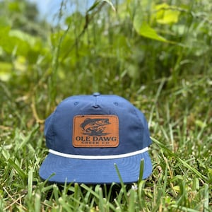 May include: A navy blue baseball cap with a brown leather patch featuring a fish graphic and the text "OLE DAWG CREEK CO." A white rope detail accents the brim. The cap is set against a backdrop of green grass and foliage.