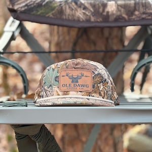 May include: Camouflage baseball cap with a brown leather patch featuring a deer silhouette and the text "OLE DAWG CREEK CO." The hat is resting on a green metal surface, with a blurred forest background.