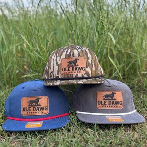 May include: Three baseball caps with a brown leather patch featuring a black silhouette of a dog and the text "Ole Dawg Creek Co.". The caps are in camouflage, blue, and gray.