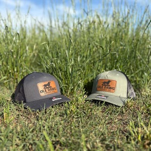 May include: Two black and green mesh trucker hats with brown leather patches. The patches feature a black silhouette of a dog and the text "OLE DAWG CREEK CO."