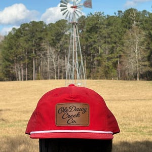 Puede incluir: Gorra de béisbol roja con detalle de cuerda blanca y un parche de cuero marrón que dice "Ole Dawg Creek Co.". La gorra se encuentra frente a un paisaje rural con un molino de viento y árboles.