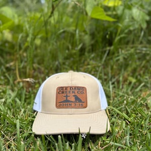 May include: A tan and white trucker hat with a rectangular patch featuring the text "OLE DAWG CREEK CO." and "JOHN 3:16". The patch also includes a cross and a dog silhouette. The hat is sitting in green grass.