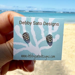 May include: A pair of seashell stud earrings on a light blue card with the text "Debby Sato Designs" and website address. The earrings are black and white, and the card has a white coral design. The background is a beach with blue water and sky.