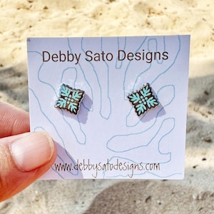 May include: A pair of square stud earrings with a turquoise and brown floral design, set against a white card with the text "Debby Sato Designs" and the website address. The earrings are held by a person.