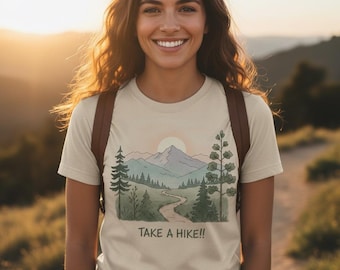 May include: Beige t-shirt with a mountain and trail graphic, and the text "TAKE A HIKE!!". The design features a winding path, trees, and a setting sun. The person is wearing a brown backpack.