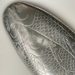 May include: A silver oval-shaped dish with an embossed design of three fish. The fish are detailed with scales and fins.