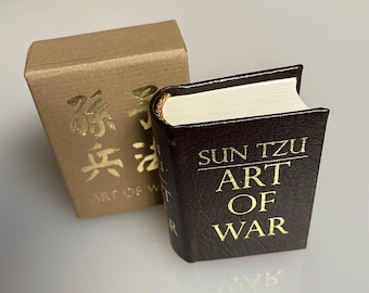May include: A miniature book with a dark brown cover and gold lettering that reads "SUN TZU ART OF WAR." The book has gold-gilded edges and is next to a small, tan box with gold text. The book is a collectible item.