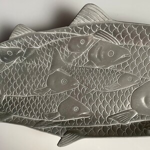 May include: A silver metal serving tray shaped like a fish with smaller fish embossed on the surface. The tray is designed to hold food.