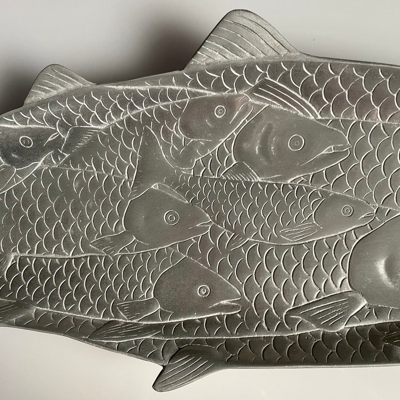May include: A silver metal serving tray shaped like a fish with smaller fish embossed on the surface. The tray is designed to hold food.