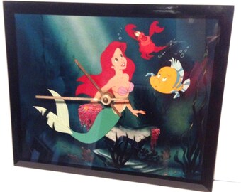 May include: A framed clock featuring a cartoon image of Ariel the mermaid with red hair, a pink seashell bra, and a green tail. The clock also shows the characters Sebastian the crab and Flounder the fish. The clock hands are brown.