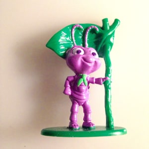 May include: A purple cartoon ant figurine holding a green leaf. The ant is smiling and has large eyes. The figurine is standing on a green base.