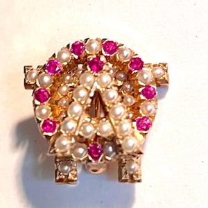 May include: A gold-toned brooch in the shape of the letter "A", encrusted with small pearls and vibrant pink gemstones. The brooch has a classic design with a pin closure on the back, suitable for adorning clothing or accessories.