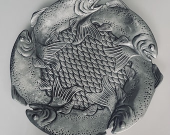 May include: A circular silver-toned metal plate featuring a relief design of four fish arranged in a circular pattern. The center of the plate displays a detailed, overlapping scale texture, while the fish bodies are textured with small dots.