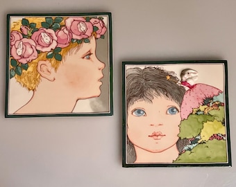 May include: Two framed ceramic tiles with illustrations of children. The first tile features a child with blonde hair wearing a flower crown. The second tile features a child with dark hair wearing a crown with a white rabbit figurine. Both tiles have a green border.