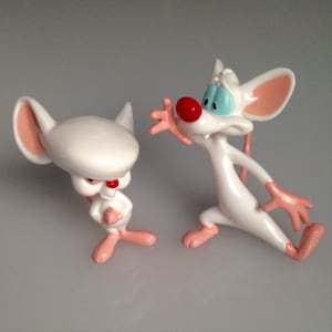May include: Two white cartoon mouse figurines, one with a red nose and a serious expression, the other with a red nose and a surprised expression. The mice have pink ears, hands, and feet. The figurines are on a gray surface.