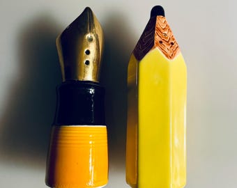 May include: A pair of ceramic salt and pepper shakers. One is shaped like a yellow pencil with a brown tip, and the other is a pen nib with gold and black accents. The pencil shaker is yellow, and the pen shaker is black and gold.