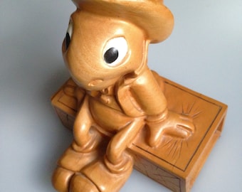 May include: A tan-colored figurine of a cartoon cricket wearing a top hat and sitting on a rectangular box. The cricket has large eyes and is dressed in a suit. The box has etched lines on the top.