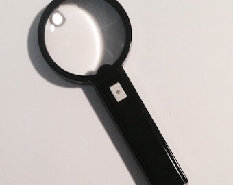 May include: A black handheld magnifying glass with a round lens and a black handle. The handle has a small white button.
