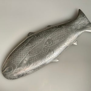 May include: A silver metal fish-shaped serving dish with an embossed design of fish swimming in a school. The dish is a decorative piece for serving food.