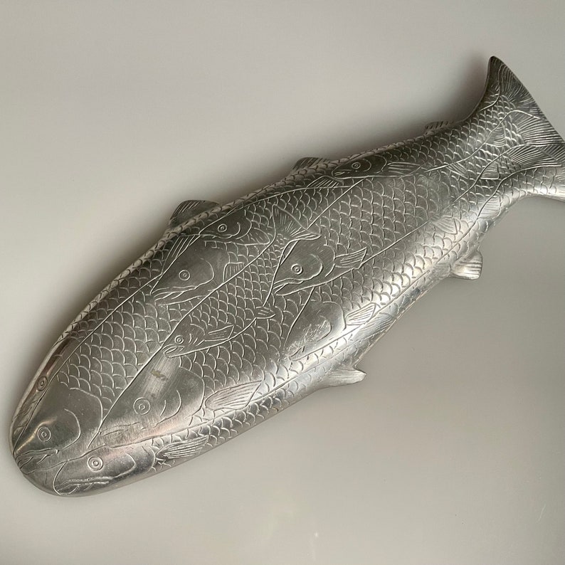 May include: A silver metal fish-shaped serving dish with an embossed design of fish swimming in a school. The dish is a decorative piece for serving food.