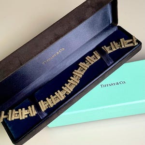 May include: A gold-toned bracelet with a geometric design, in a Tiffany & Co. box. The box is brown with a blue interior and the Tiffany & Co. logo is printed on the box.