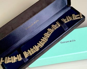 May include: A gold-toned bracelet with a geometric design, in a Tiffany & Co. box. The box is brown with a blue interior and the Tiffany & Co. logo is printed on the box.