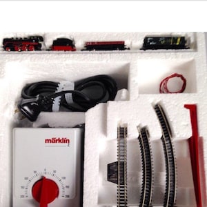 May include: A model train set featuring a red and black locomotive, freight cars, and track pieces. The set includes a power supply unit with a red dial and the brand name "märklin" printed on it. The set is packaged in a white foam insert.