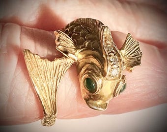 May include: A gold-colored fish ring with green gemstone eyes and small clear stones. The ring features a detailed fish design with fins and a tail that wraps around the finger. The ring is a unique piece of jewelry.