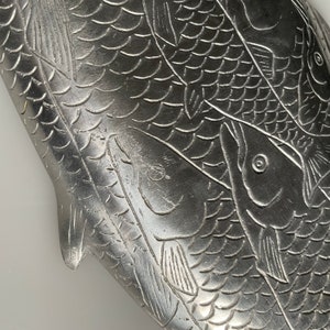 May include: A silver metal tray with an embossed design of three fish. The fish are arranged in a overlapping pattern, with their scales and fins visible. The tray has a smooth, polished surface.