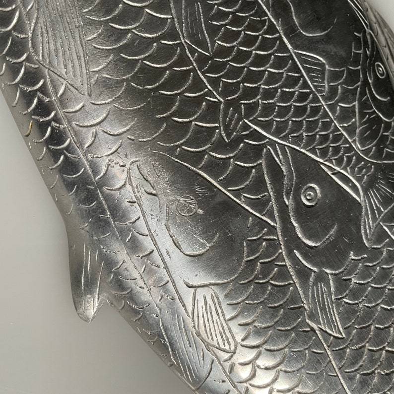 May include: A silver metal tray with an embossed design of three fish. The fish are arranged in a overlapping pattern, with their scales and fins visible. The tray has a smooth, polished surface.